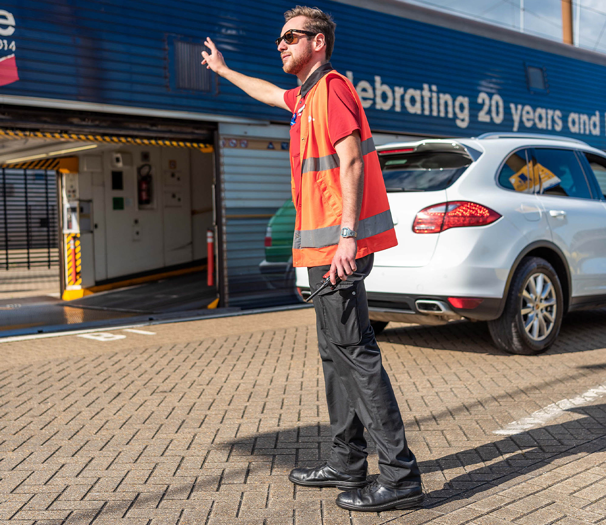 Man with brown hair and beard wearing sunglasses a bright orange vest and dark trousers signalling with his arms to cars to board them on a train